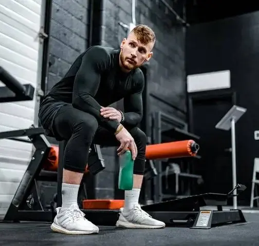 A fit man in black athletic wear sitting on a gym bench, holding a water bottle, representing strength, fitness, and gym dedication.