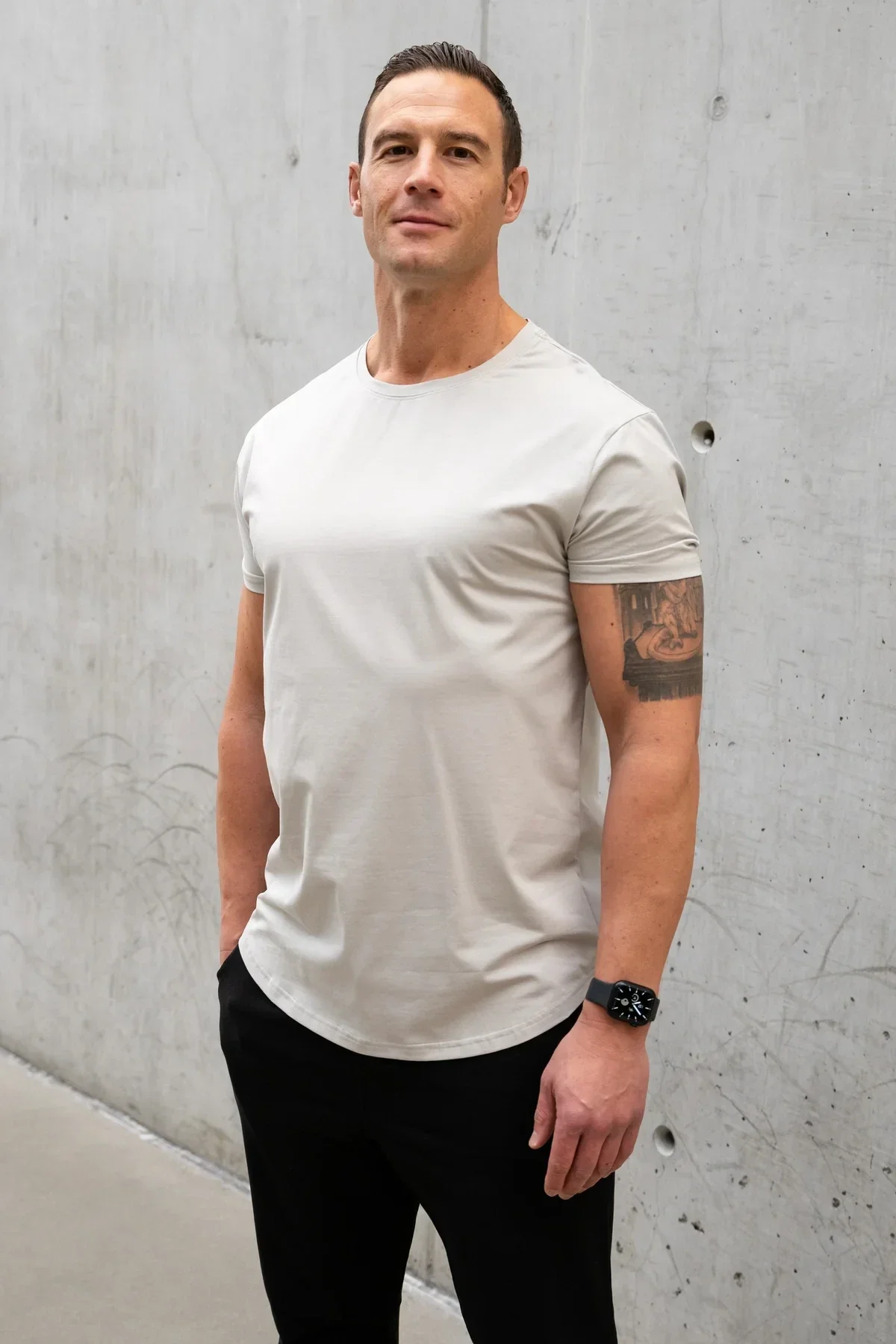 Man wearing a light grey short-sleeved T-shirt and black pants, standing confidently against a grey concrete wall.