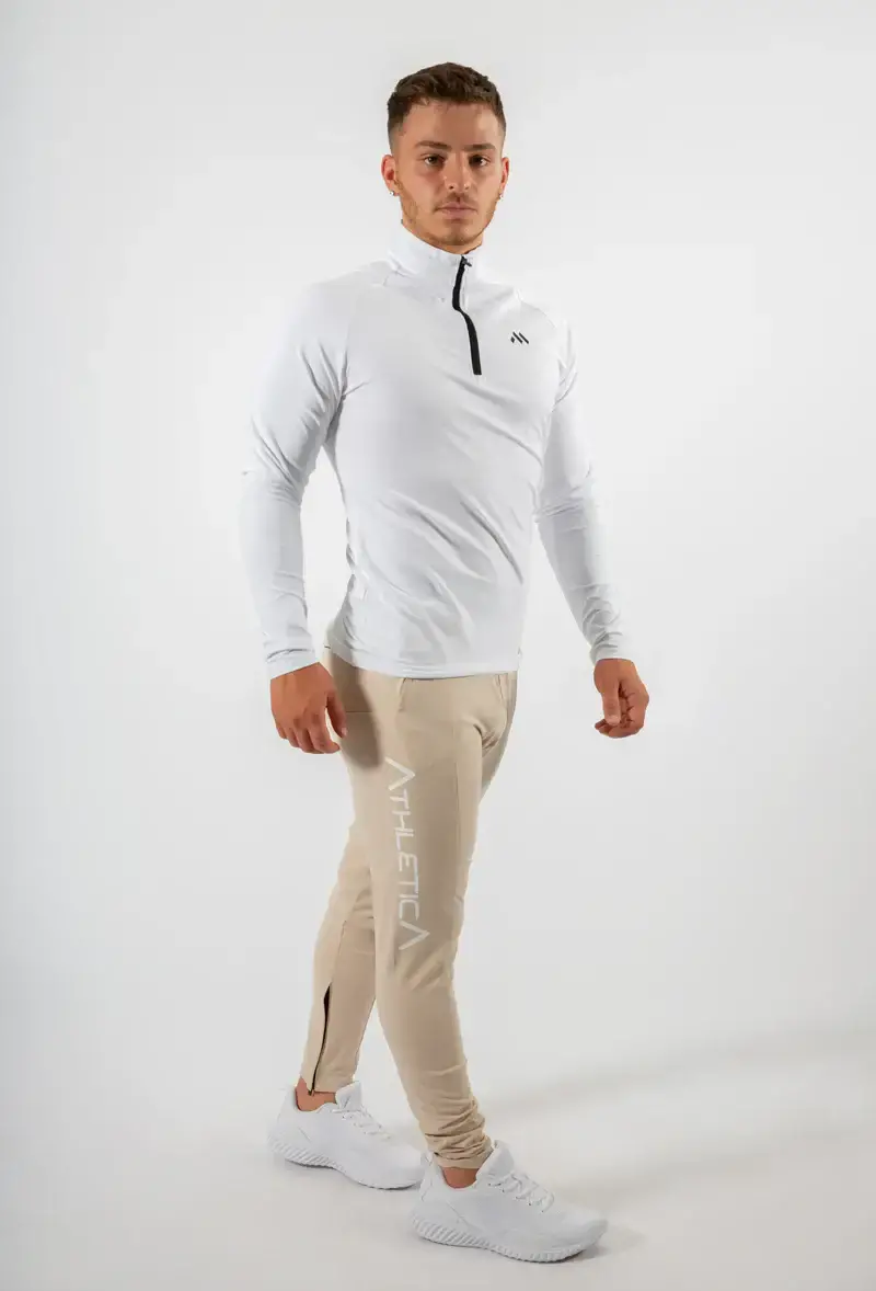 A man modeling Athletica Athleisure men’s wear, featuring a white zip-up top and beige joggers.