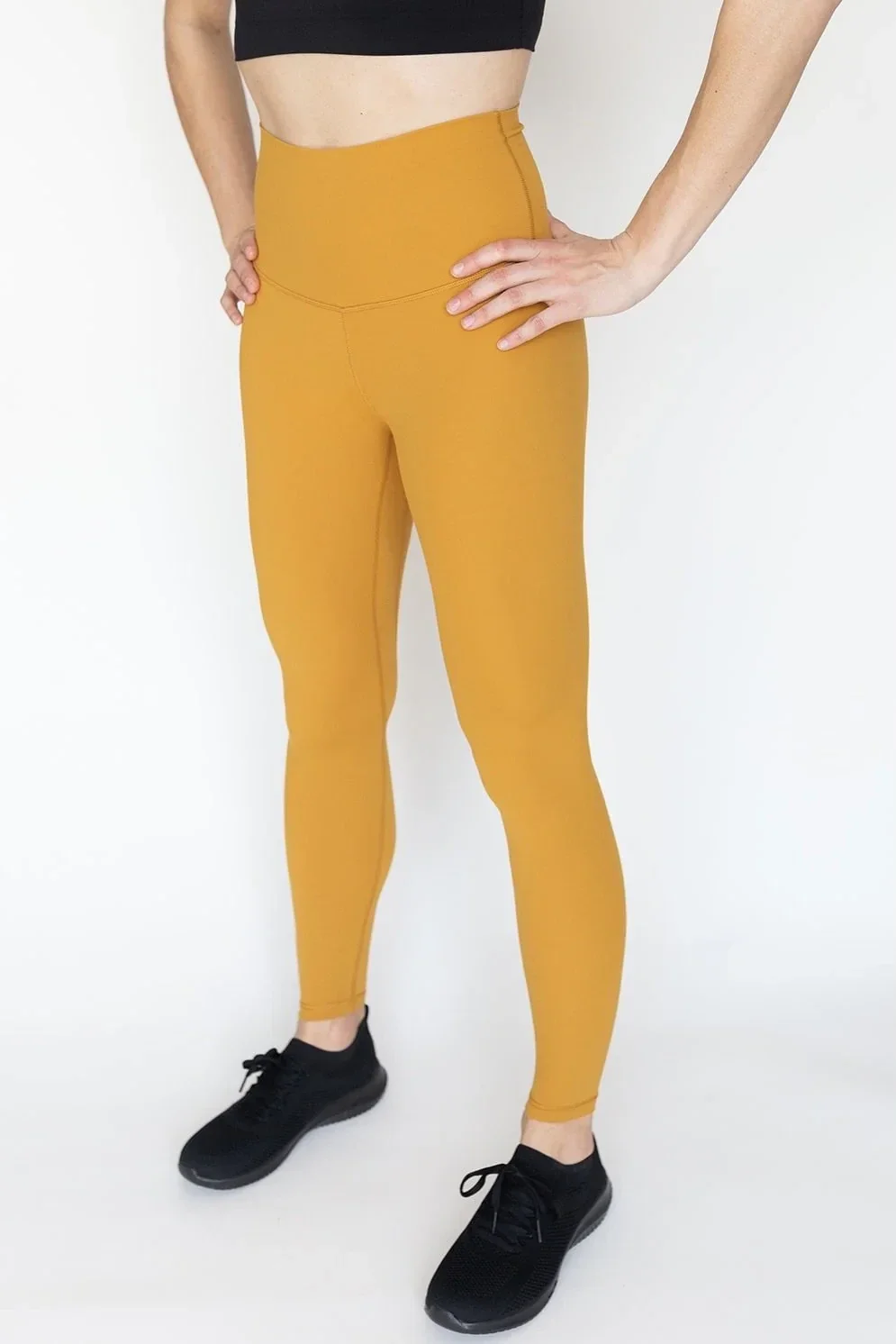 Woman wearing mustard yellow high-waisted leggings with black sneakers against a plain white background.