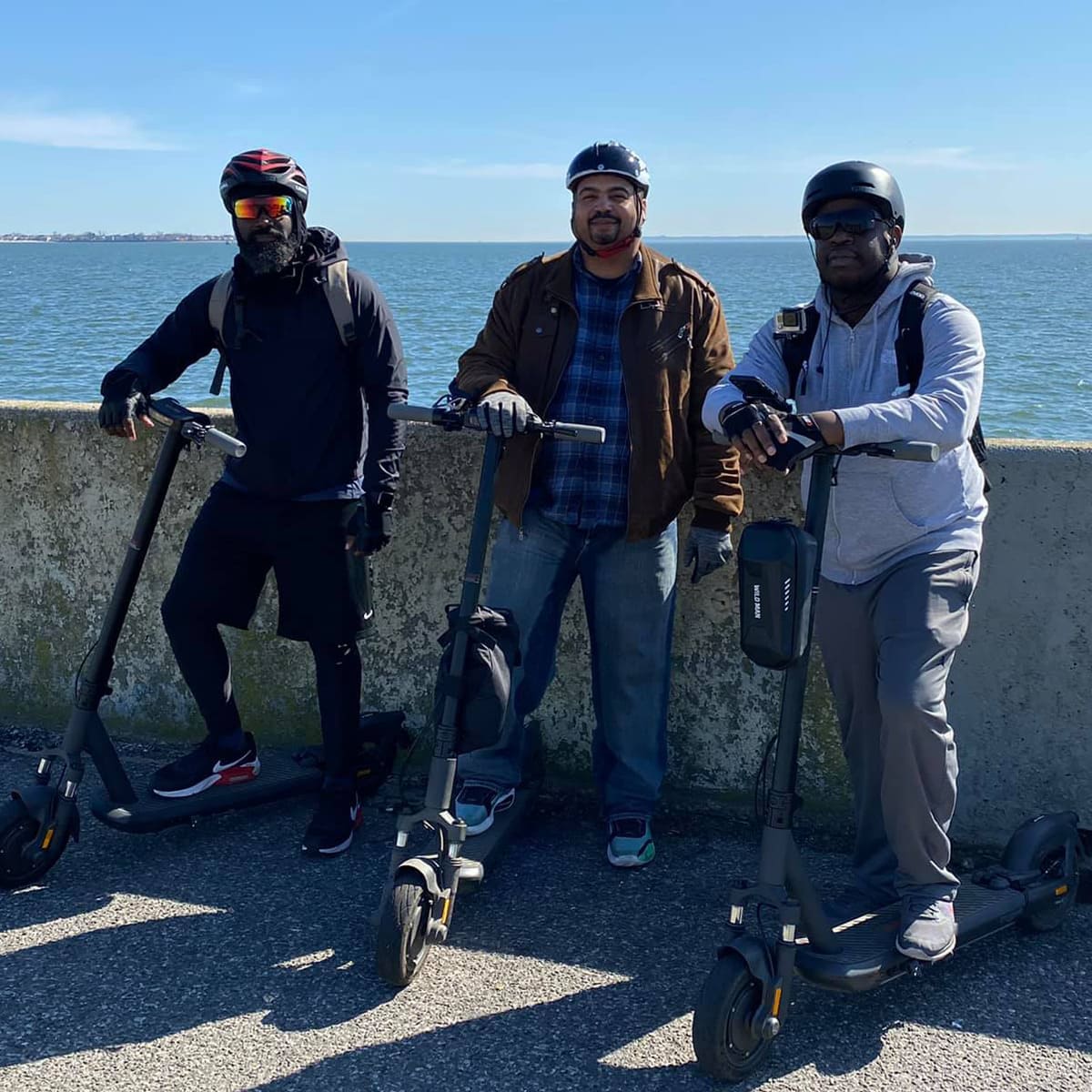 Three riders posing with InMotion S1F electric scooters near a coastal area on a sunny day.