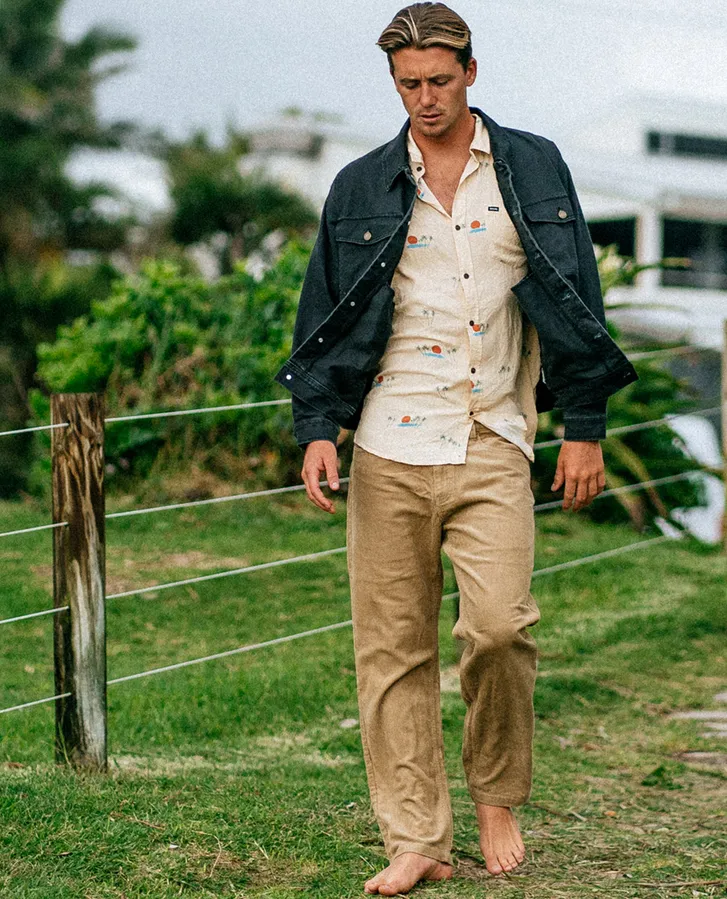 Model wearing Rip Curl tan corduroy pants paired with a patterned shirt and jacket, walking outdoors barefoot.