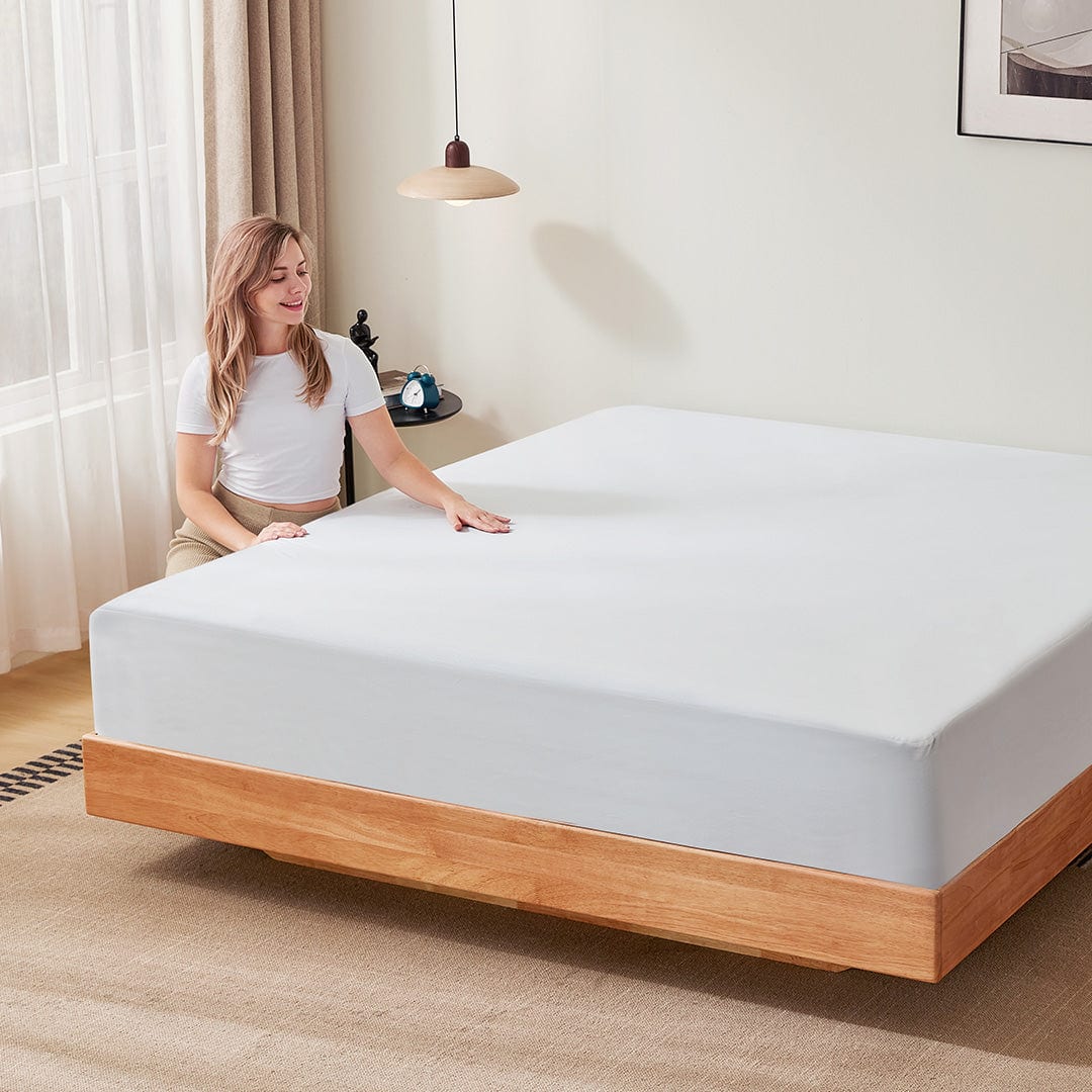 Woman smiling and placing her hand on a perfectly made bed featuring a Newentor mattress protector in a bright, modern bedroom. Enhance your health and fitness journey with quality sleep at www.waiteandsea.com.au.