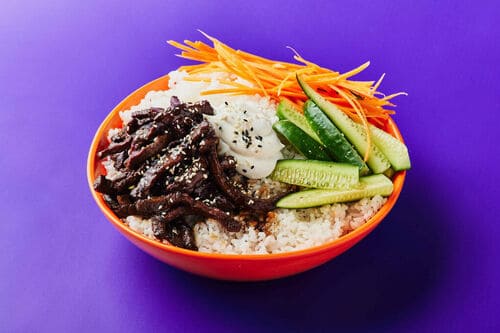 A vibrant sushi bowl featuring teriyaki beef, rice, fresh cucumber, carrot ribbons, and a drizzle of creamy dressing.