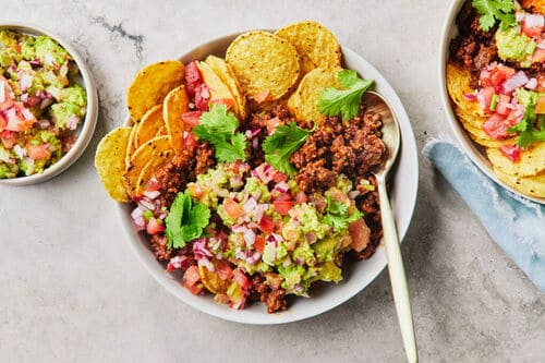 A bowl of Tex Mex beef nachos topped with fresh guacamole, salsa, and coriander, served with crunchy tortilla chips.