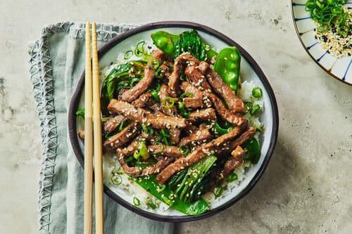 Garlic pepper beef stir fry served with fresh snow peas, bok choy, and steamed rice, garnished with sesame seeds and spring onions.