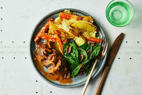 A hearty plate of roast vegetables, sautéed spinach, and tender steak topped with rich gravy, served with a refreshing green drink.