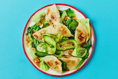 A plate of pan-fried pork dumplings served with fresh sesame greens and cucumber ribbons on a vibrant blue background.