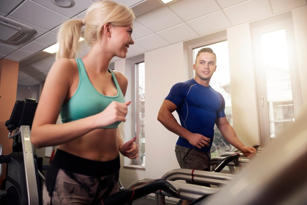 A fit man and woman jogging on treadmills in a bright gym, smiling at each other. Keywords: health and fitness, cardio workout, treadmill running, endurance training, www.waiteandsea.com.au