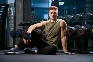 Young male athlete in a gym sitting in front of a dumbbell rack, focused on muscle-building workouts for skinny guys. Strength training and health and fitness. www.waiteandsea.com.au